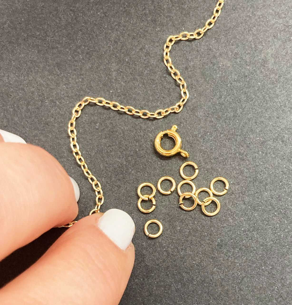 Gold chain and jump rings on a gray surface with a hand holding the chain.